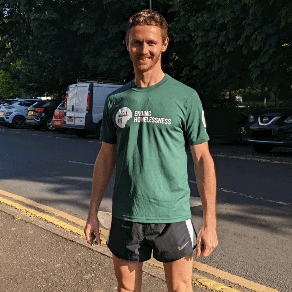 A man in a green tshirt and black running shorts stands on the pavement smiling