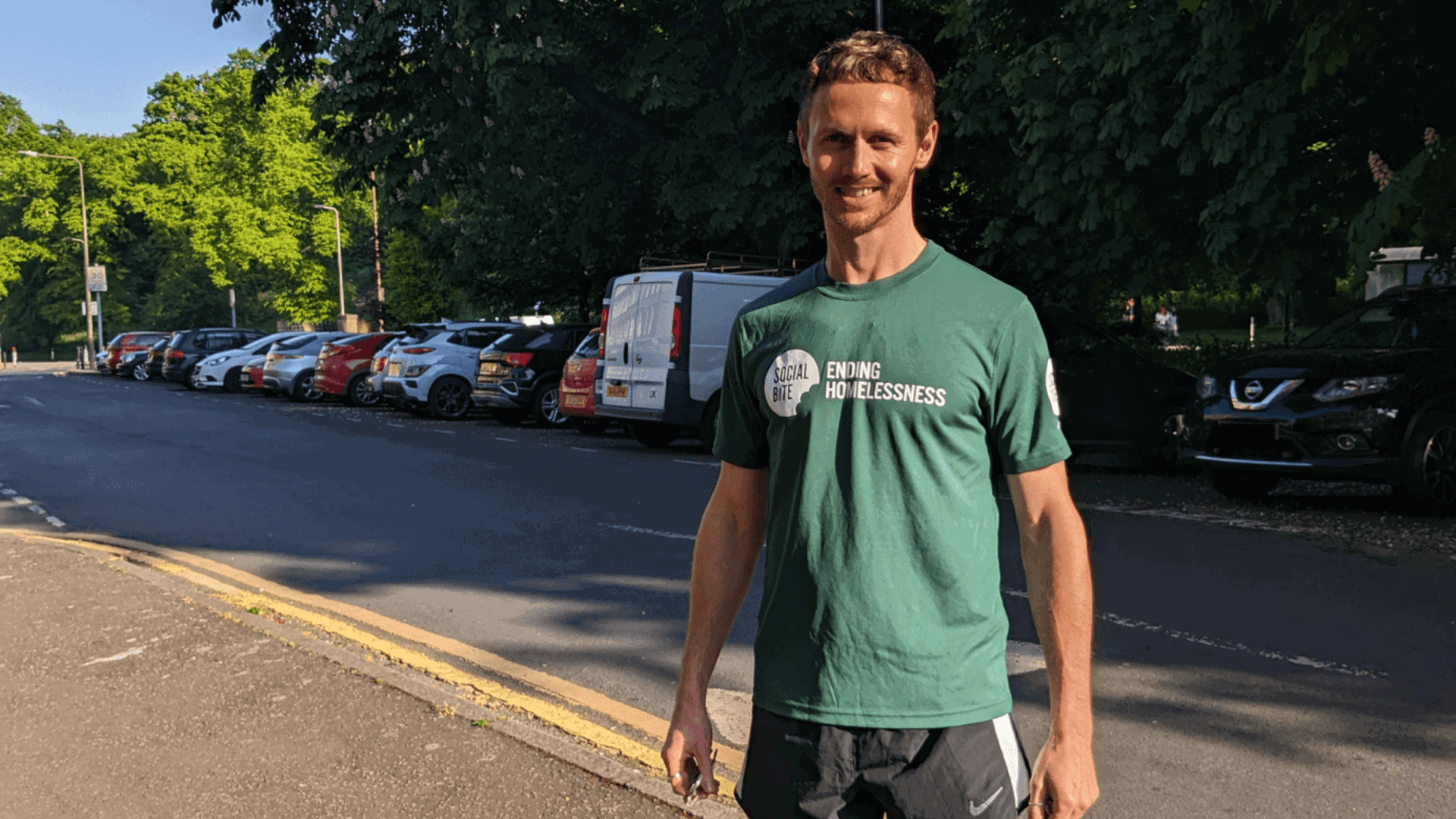 A man in a green tshirt and black running shorts stands on the pavement smiling