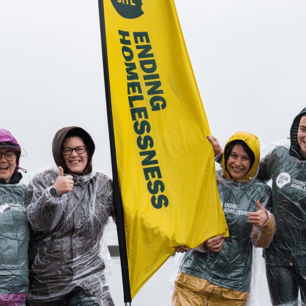 Four women in raincoats and clear ponchos smile and give thumbs up in very wet weather. They have a tall yellow banner between them that reads 'ending homelessness'