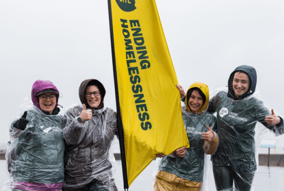 Four women in raincoats and clear ponchos smile and give thumbs up in very wet weather. They have a tall yellow banner between them that reads 'ending homelessness'