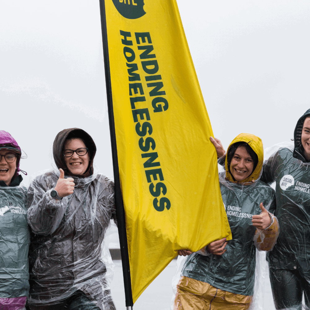 Four women in raincoats and clear ponchos smile and give thumbs up in very wet weather. They have a tall yellow banner between them that reads 'ending homelessness'