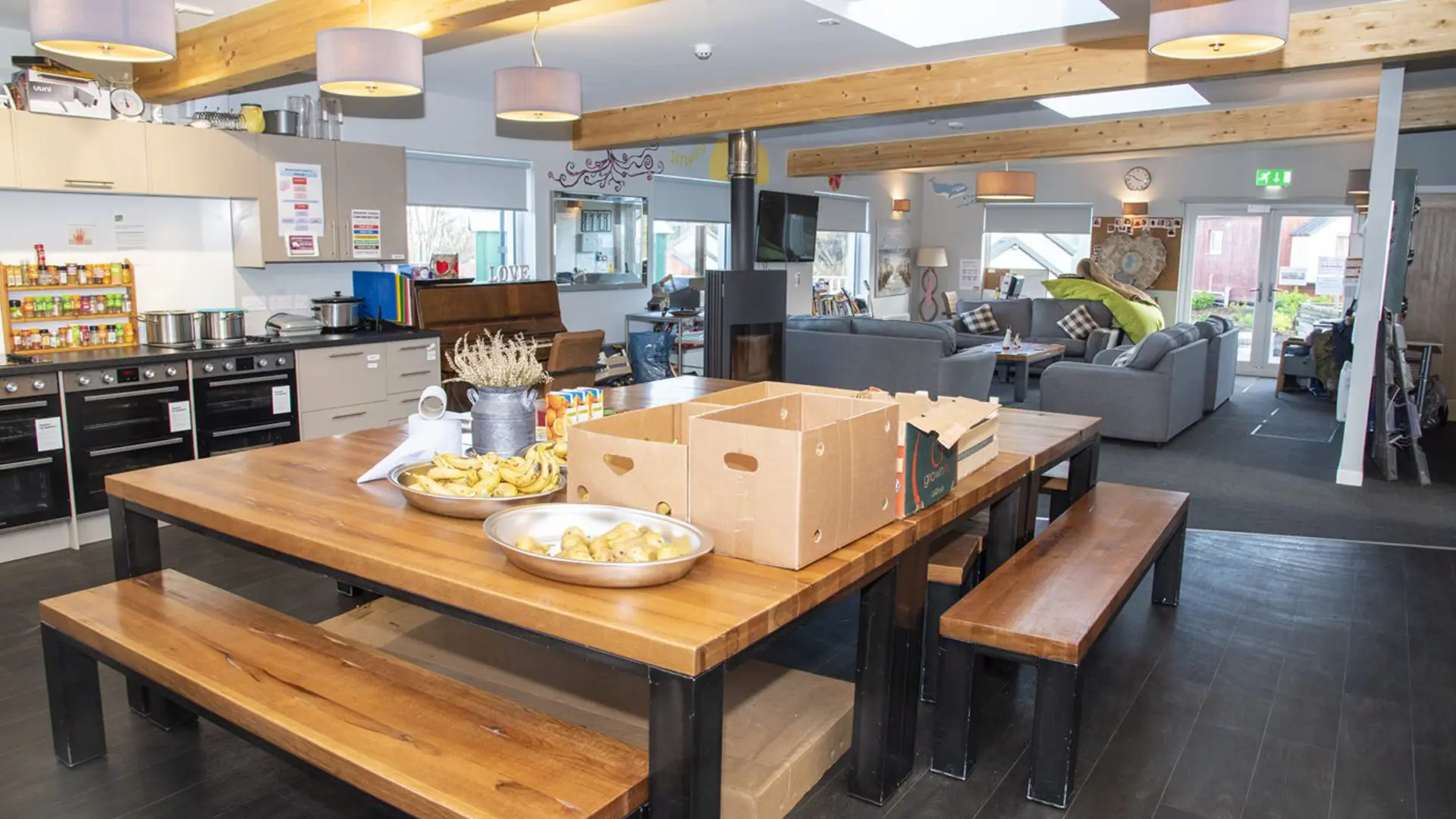 A huge communal kitchen dining table with boxes and snacks on it, looking on to a living room
