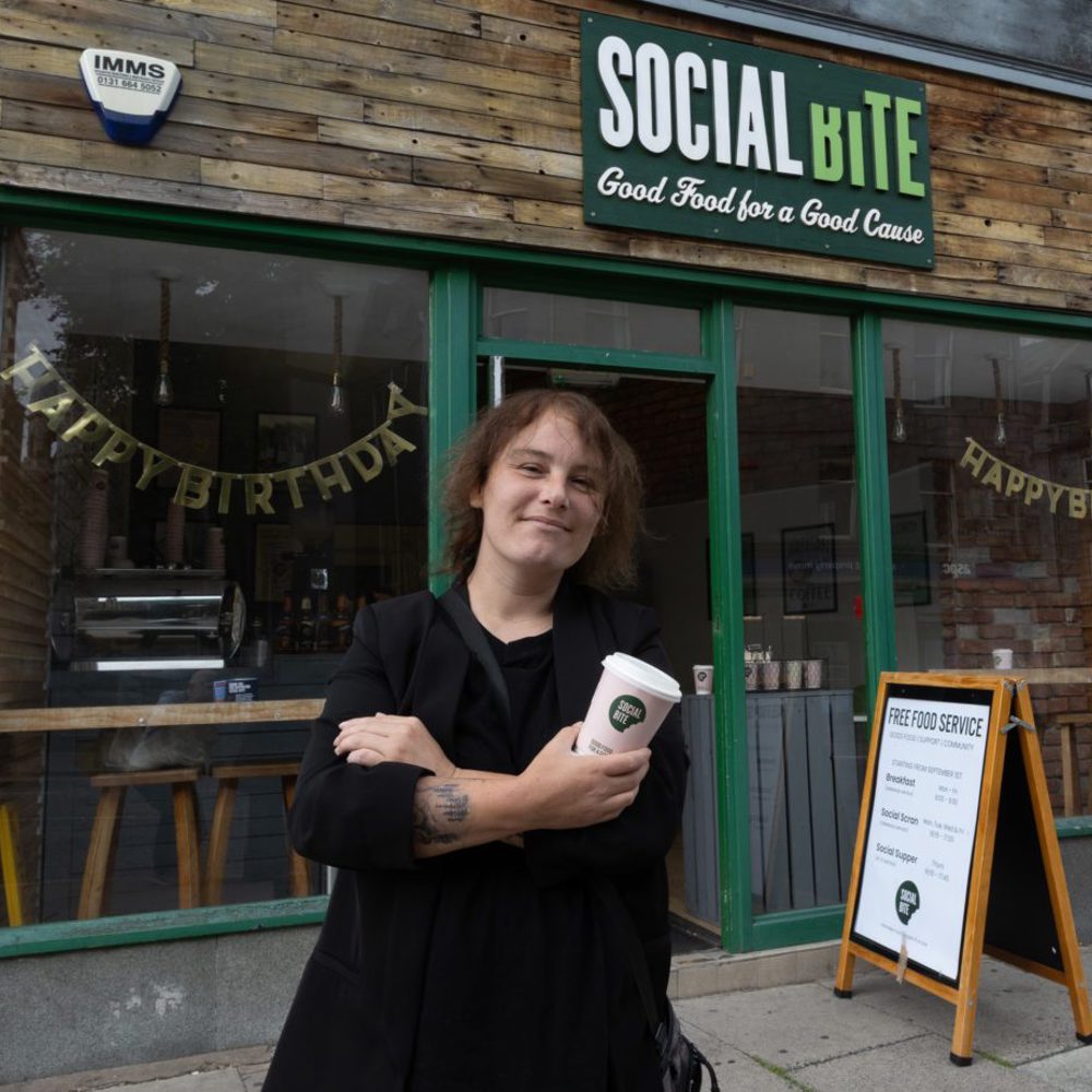 A woman holding a takeaway coffee cup standing with arms crossed outside a shop called Social BIte