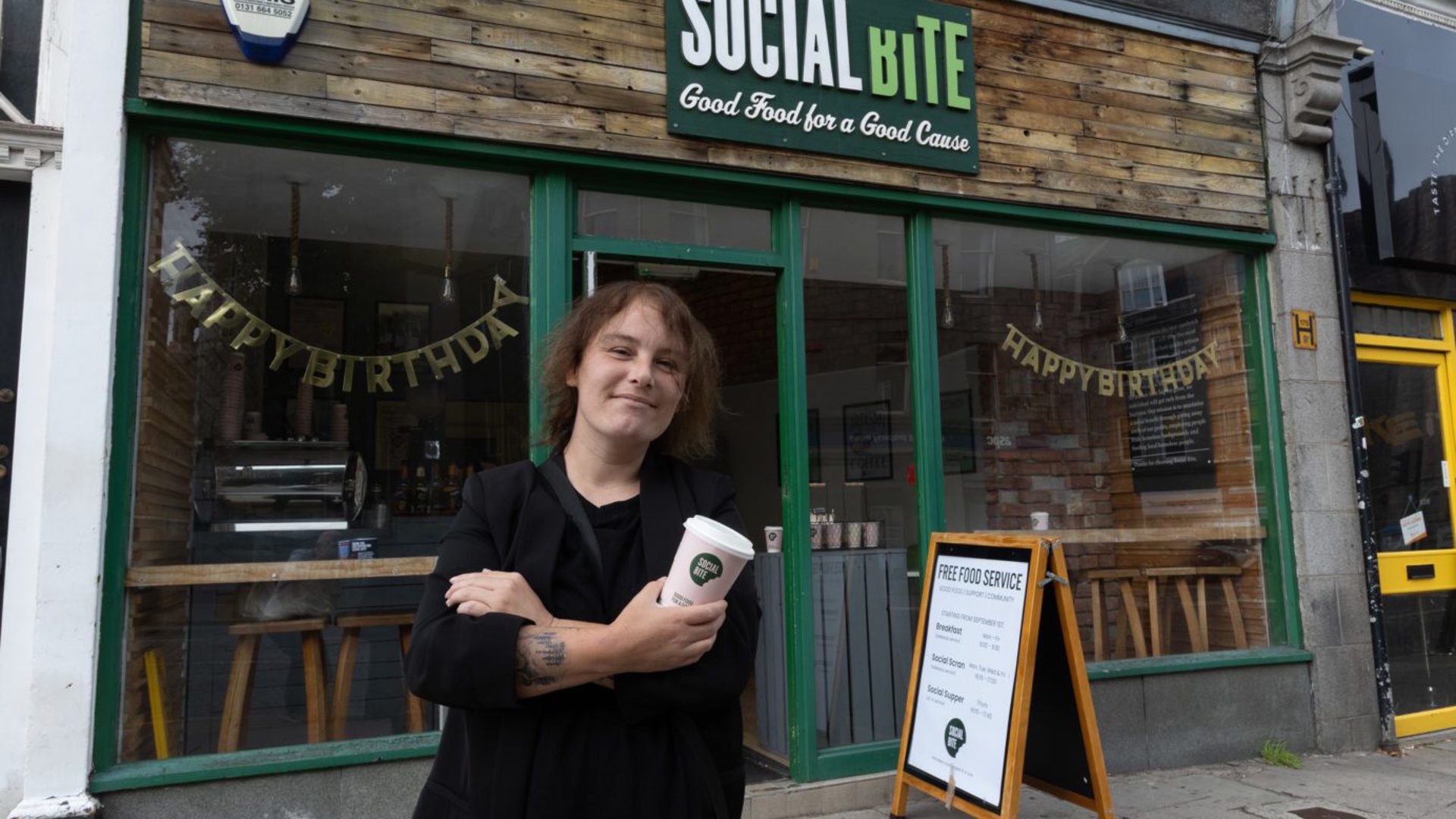 A woman holding a takeaway coffee cup standing with arms crossed outside a shop called Social BIte