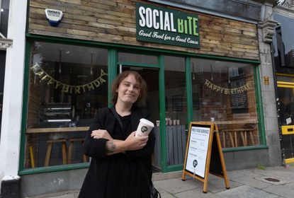 A woman holding a takeaway coffee cup standing with arms crossed outside a shop called Social BIte