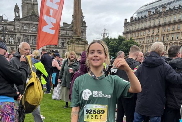 A young woman in a Social Bite tshirt poses with her medal in front of a crowd of people after completing a 10k race