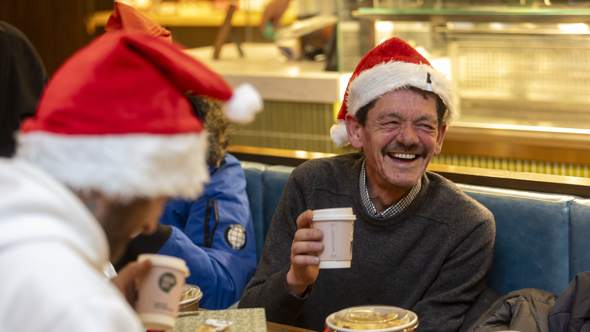 People smiling and enjoying hot food and hot drinks with santa hats on and crackers on the table. 