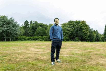 A man in a blue sweatshirt stands in a field with trees