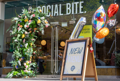 Birthday balloons on a shop A board outside a glass-fronted shop called Social Bite
