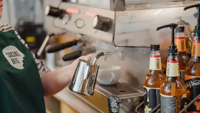 A woman in a green apron steaming milk at an espresso machine