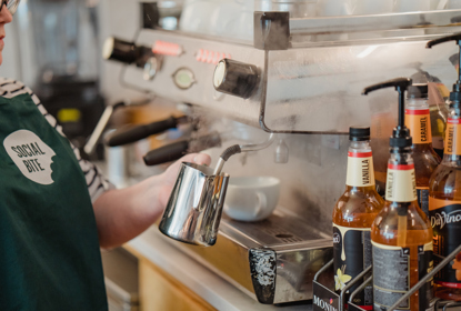 A woman in a green apron steaming milk at an espresso machine