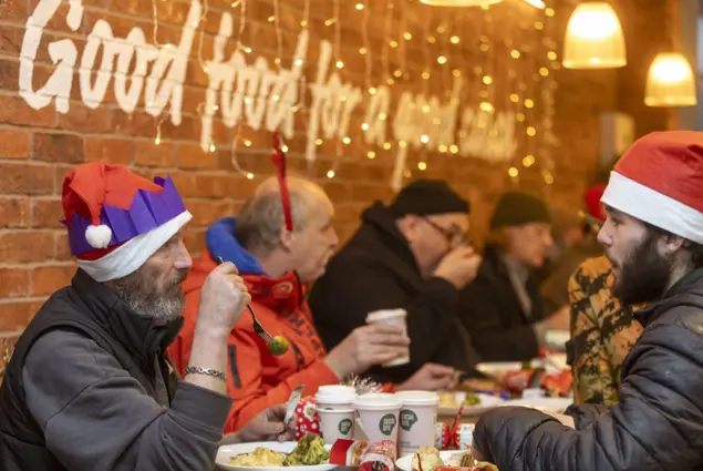 Men in Santa hats and paper crowns eat a meal at a long table, with sparkling lights and Christmas crackers around them