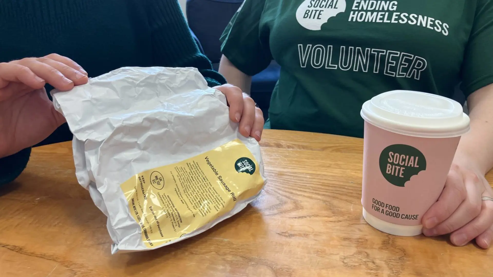Two people sit together with a coffee. One wears a green tshirt that says volunteer.