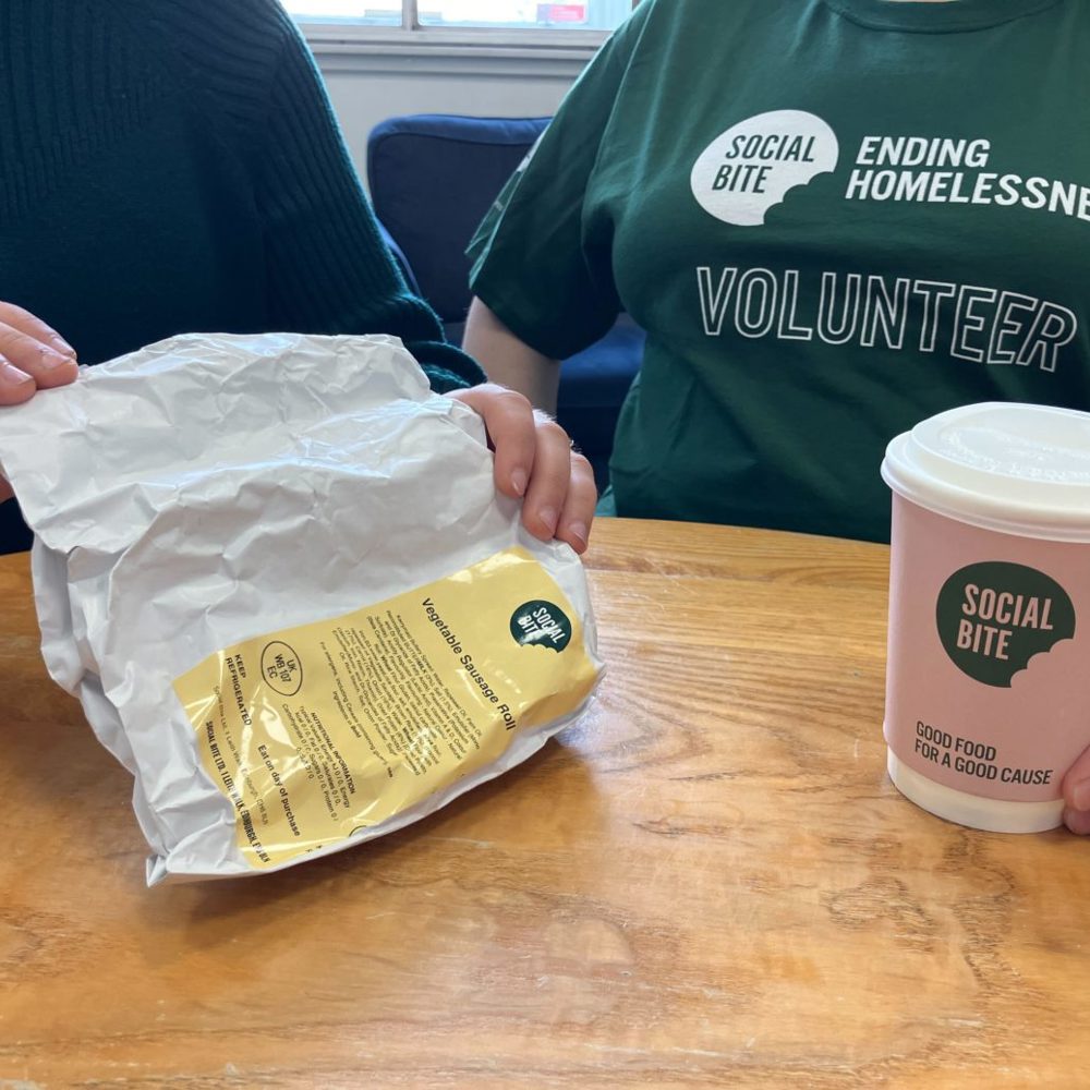 Two people sit together with a coffee. One wears a green tshirt that says volunteer.