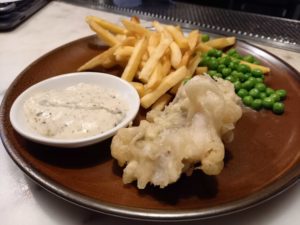 A brown plate with battered fish, chips, peas and a bowl of tartare sauce
