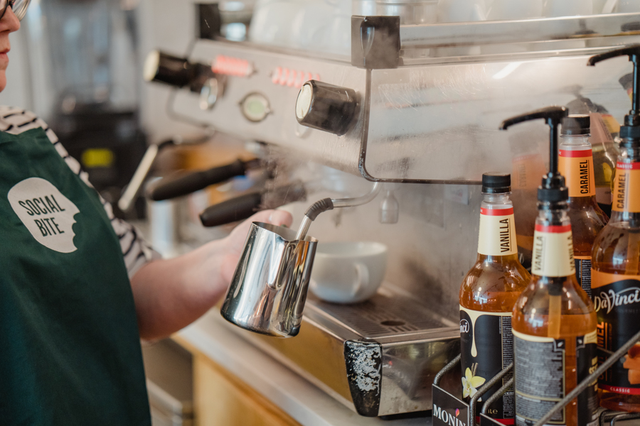 A woman in a green apron steaming milk at an espresso machine