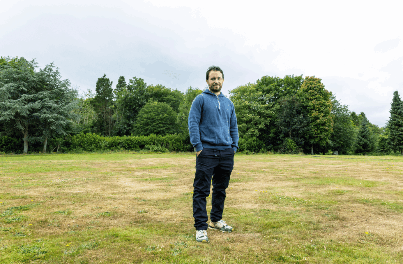 A man in a blue sweatshirt stands in a field with trees