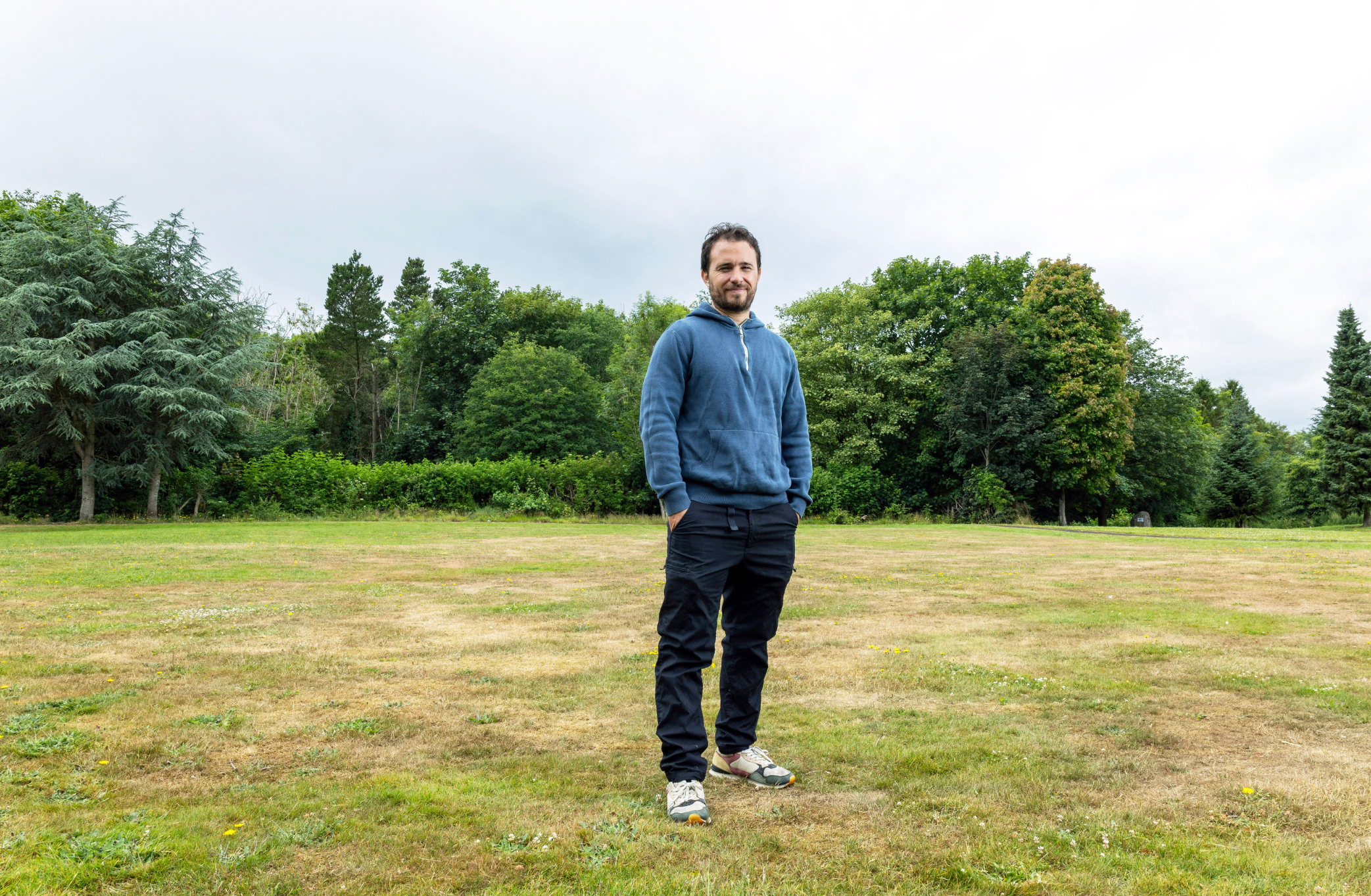 A man in a blue sweatshirt stands in a field with trees